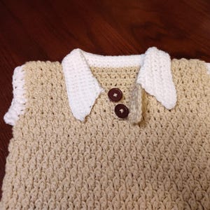 A handmade crochet vest in a light tan color with a white collar and sleeve trim. Two brown buttons fasten the collar. The textured stitch creates a cozy, classic look. This vest is perfect for a baby or toddler.