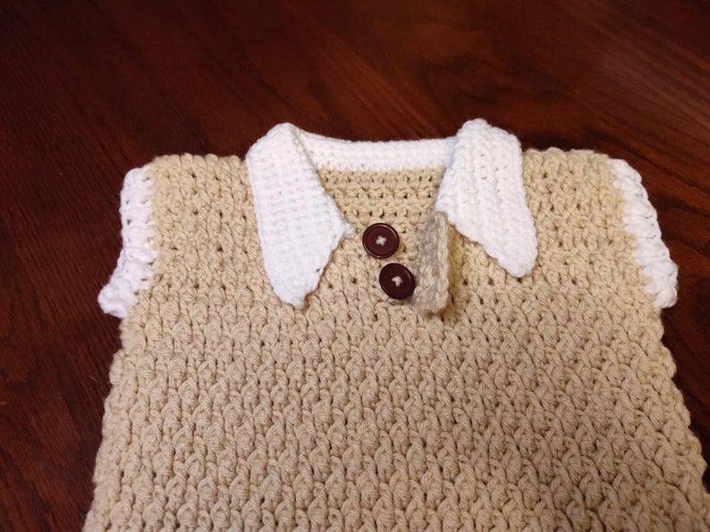 A handmade crochet vest in a light tan color with a white collar and sleeve trim. Two brown buttons fasten the collar. The textured stitch creates a cozy, classic look. This vest is perfect for a baby or toddler.