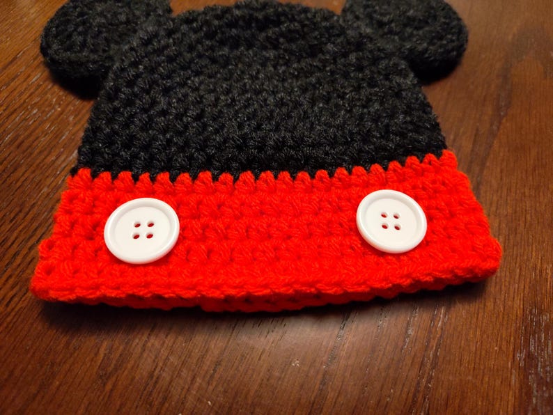 A black and red crocheted hat featuring two round black ears on top and two large white buttons attached to the front red section. The hat is constructed with a textured yarn stitch pattern and has a distinct color-blocked design.