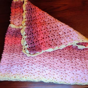 A handmade crochet blanket in shades of pink and peach, with a cream-colored border. The blanket is folded, showcasing the intricate stitch pattern and color gradient. The soft, textured blanket is likely made of yarn.