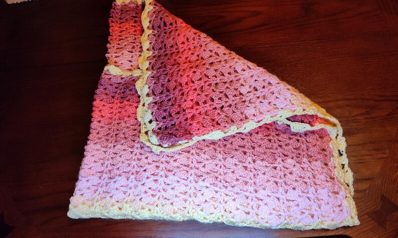 A handmade crochet blanket in shades of pink and peach, with a cream-colored border. The blanket is folded, showcasing the intricate stitch pattern and color gradient. The soft, textured blanket is likely made of yarn.