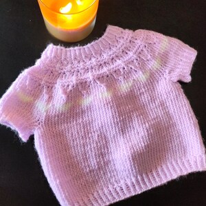 A short-sleeved, light purple knitted sweater featuring a ribbed crew neckline, a textured yoke with a subtle white horizontal stripe pattern, and a ribbed hem. The garment is crafted with a soft, fuzzy yarn texture throughout.