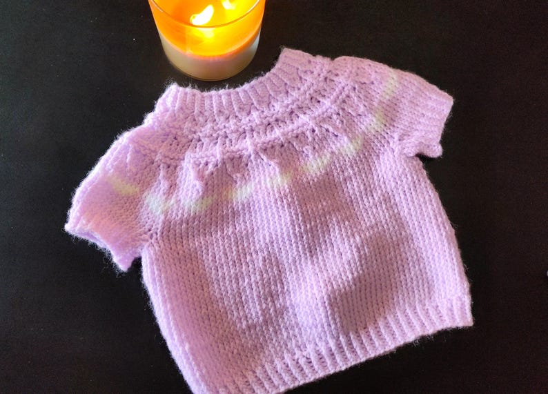 A short-sleeved, light purple knitted sweater featuring a ribbed crew neckline, a textured yoke with a subtle white horizontal stripe pattern, and a ribbed hem. The garment is crafted with a soft, fuzzy yarn texture throughout.