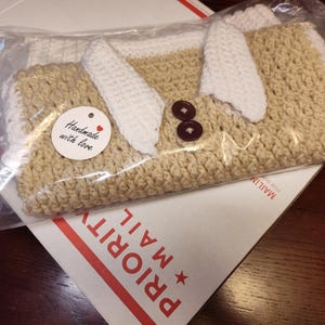 A handmade crochet sweater vest in a clear plastic bag. The vest is tan with white accents. Two brown buttons are visible. A small tag reads "Handmade with love."