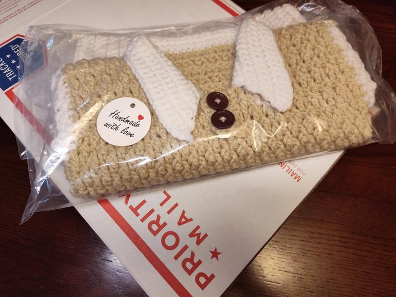 A handmade crochet sweater vest in a clear plastic bag. The vest is tan with white accents. Two brown buttons are visible. A small tag reads "Handmade with love."