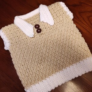 A handmade crochet vest in tan with white trim and collar. The vest features two brown buttons and a textured stitch pattern. The collar and trim are a contrasting white color, creating a classic look.