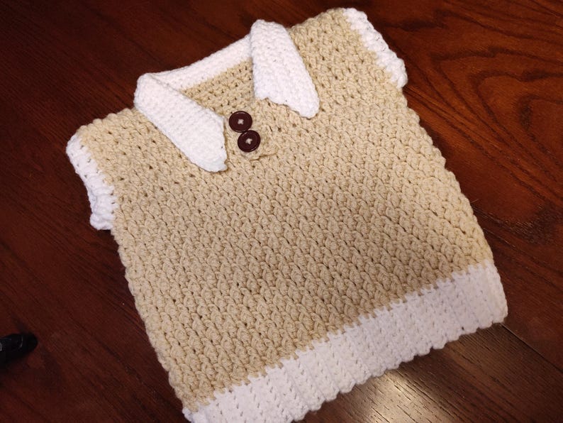 A handmade crochet vest in tan with white trim and collar. The vest features two brown buttons and a textured stitch pattern. The collar and trim are a contrasting white color, creating a classic look.
