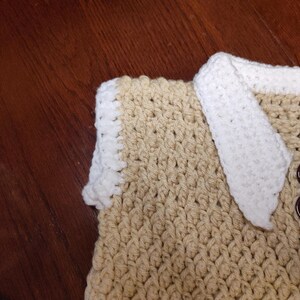 Close-up of a handmade crochet item in tan and white. The item features a textured stitch pattern, with a white border and accents. Two brown buttons are visible. The item is likely a sweater vest.