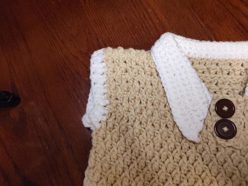 Close-up of a handmade crochet item in tan and white. The item features a textured stitch pattern, with a white border and accents. Two brown buttons are visible. The item is likely a sweater vest.