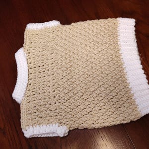 A handmade crochet garment in a neutral beige color with white trim. The crochet stitch creates a textured pattern. The garment appears to be a child's vest.