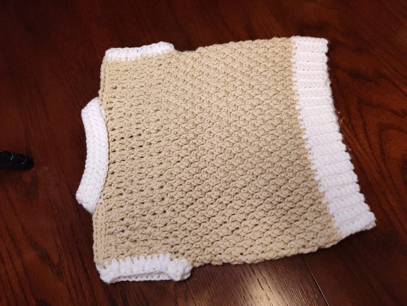 A handmade crochet garment in a neutral beige color with white trim. The crochet stitch creates a textured pattern. The garment appears to be a child's vest.