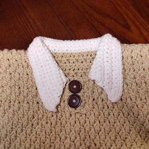 Crocheted beige sweater vest with white trim and accents. Features two brown buttons and white crocheted collar. The sweater has a textured pattern and is handmade.