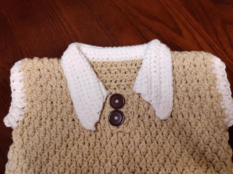 Crocheted beige sweater vest with white trim and accents. Features two brown buttons and white crocheted collar. The sweater has a textured pattern and is handmade.