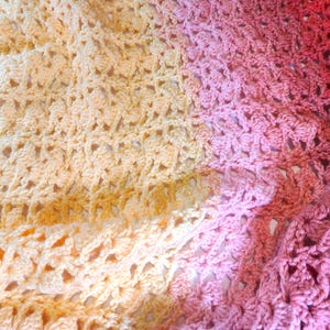 A handmade crochet blanket in shades of yellows, pinks, reds and purples. The blanket features a textured pattern and a scalloped edge.