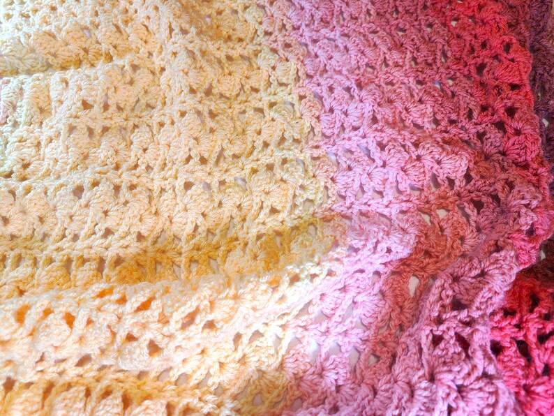 A handmade crochet blanket in shades of yellows, pinks, reds and purples. The blanket features a textured pattern and a scalloped edge.