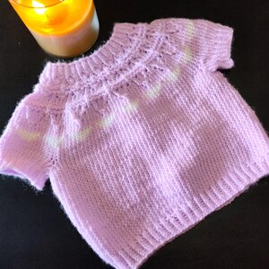 A short-sleeved, light purple knitted sweater featuring a textured yoke with subtle white accents. The garment has a ribbed crew neckline, ribbed sleeve cuffs, and a ribbed hem, showcasing a consistent knit pattern throughout the body.