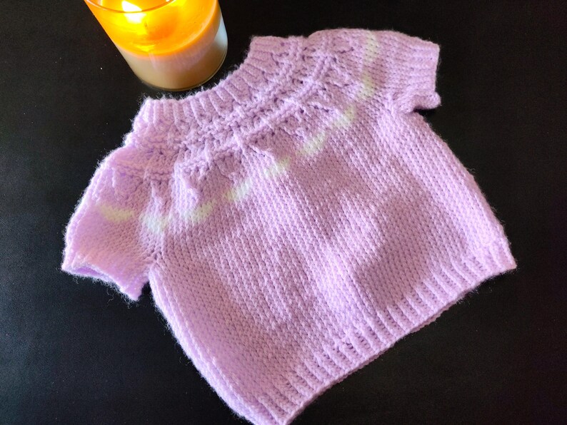 A short-sleeved, light purple knitted sweater featuring a textured yoke with subtle white accents. The garment has a ribbed crew neckline, ribbed sleeve cuffs, and a ribbed hem, showcasing a consistent knit pattern throughout the body.