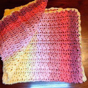 A handmade crochet blanket in shades of yellow, pink, and purple. The blanket features a textured pattern and a scalloped edge. The blanket is folded on a dark wooden surface.