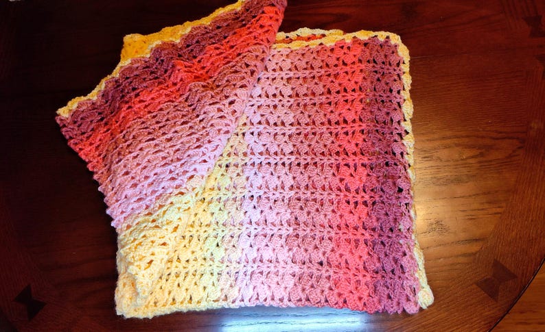 A handmade crochet blanket in shades of yellow, pink, and purple. The blanket features a textured pattern and a scalloped edge. The blanket is folded on a dark wooden surface.