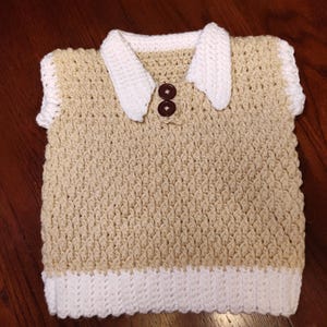 A handmade, beige and white crochet vest with a white collar and two brown buttons. The vest has a textured stitch pattern and a white border at the bottom and around the sleeves. This item is suitable for a baby or toddler.