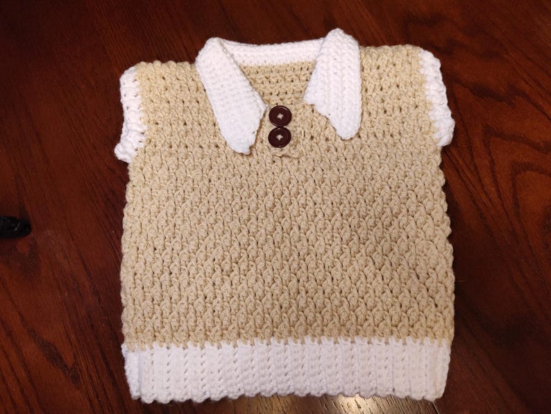 A handmade, beige and white crochet vest with a white collar and two brown buttons. The vest has a textured stitch pattern and a white border at the bottom and around the sleeves. This item is suitable for a baby or toddler.