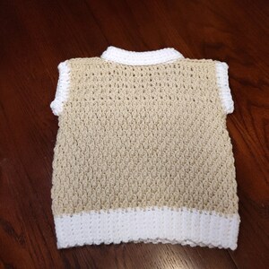 A handmade crochet vest in a neutral beige color with white trim on the collar, sleeves, and bottom hem. The vest has a textured stitch pattern and a classic, sleeveless design, suitable for layering.