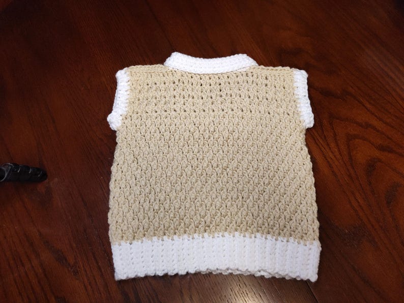 A handmade crochet vest in a neutral beige color with white trim on the collar, sleeves, and bottom hem. The vest has a textured stitch pattern and a classic, sleeveless design, suitable for layering.