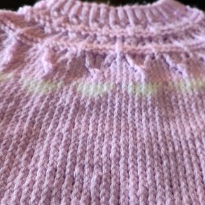 A light purple knitted sweater featuring a ribbed mock neck collar and a yoke with a decorative textured pattern. The design includes a horizontal band of white yarn integrated into the knit, creating a subtle contrast against the lavender.