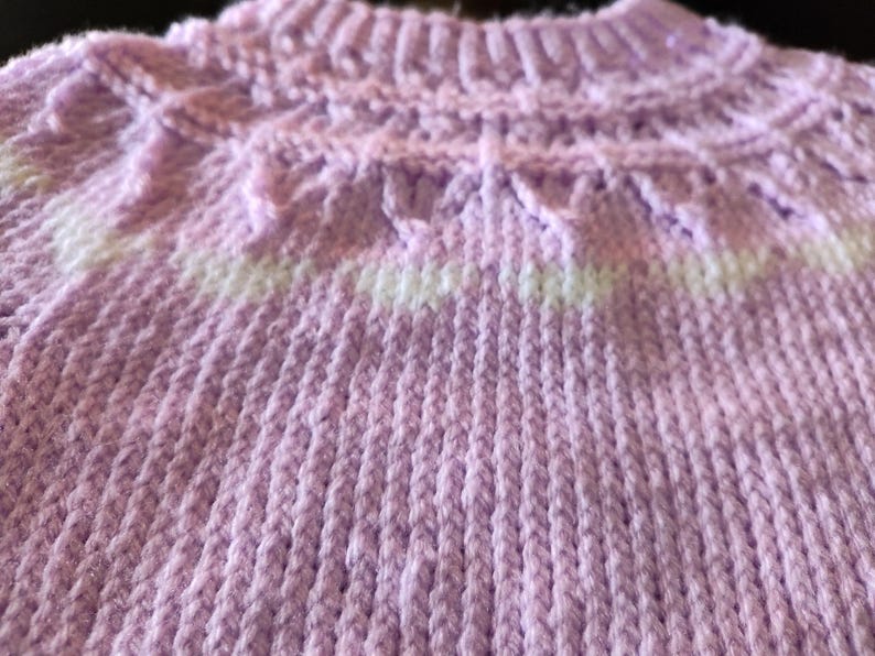 A light purple knitted sweater featuring a ribbed mock neck collar and a yoke with a decorative textured pattern. The design includes a horizontal band of white yarn integrated into the knit, creating a subtle contrast against the lavender.