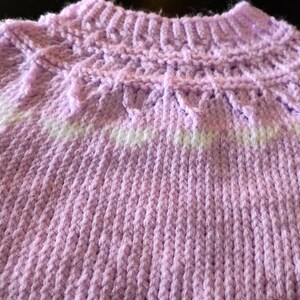 A hand-knitted sweater featuring a light lavender yarn base with a ribbed collar and a decorative yoke pattern. The yoke incorporates a horizontal band of white yarn, creating a subtle contrast against the soft purple texture of the garment.