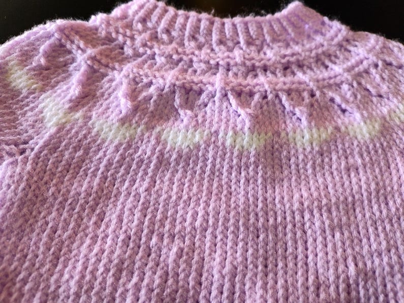 A hand-knitted sweater featuring a light lavender yarn base with a ribbed collar and a decorative yoke pattern. The yoke incorporates a horizontal band of white yarn, creating a subtle contrast against the soft purple texture of the garment.