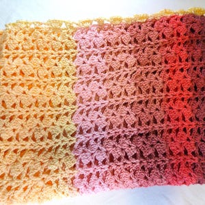 A handmade crochet blanket in shades of yellows, pinks, reds and purples. The blanket features a textured pattern and a scalloped edge.