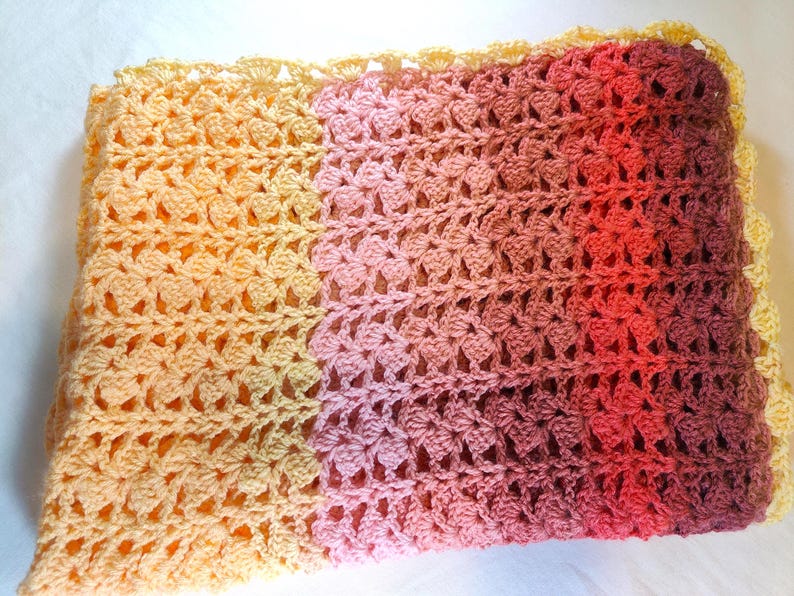 A handmade crochet blanket in shades of yellows, pinks, reds and purples. The blanket features a textured pattern and a scalloped edge.