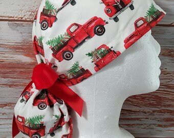 Könnte beinhalten: Eine weiße OP-Haube mit einem sich wiederholenden Muster aus roten Oldtimer-Trucks, die Weihnachtsbäume transportieren. Die Haube hat eine rote Schleife und ist zum Tragen auf dem Kopf bestimmt. Das Design ist festlich und saisonal.