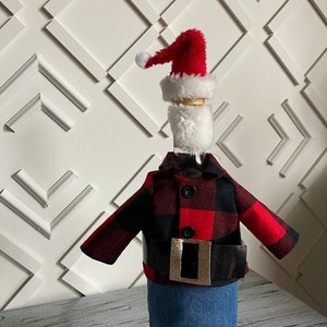 Lumberjack  Santa theme wine bottle cover showing black and red buffalo plaid jacket, blue denim bottoms, red Santa hat,  white beard and thick black belt with gold buckle to complete the look