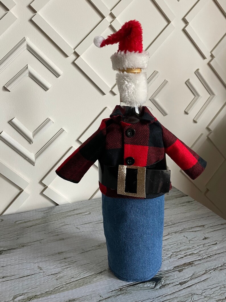 Lumberjack  Santa theme wine bottle cover showing black and red buffalo plaid jacket, blue denim bottoms, red Santa hat,  white beard and thick black belt with gold buckle to complete the look