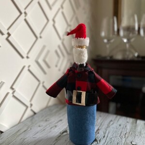 Lumberjack  Santa theme wine bottle cover showing black and red buffalo plaid jacket, blue denim bottoms, red Santa hat,  white beard and thick black belt with gold buckle to complete the look