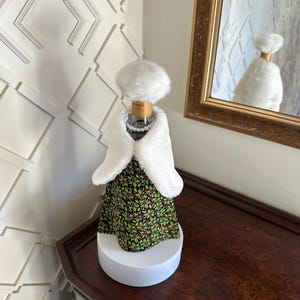 May include: A white faux fur bottle cover with a white faux fur hat and a green and red floral print dress. The bottle cover is on a white pedestal.