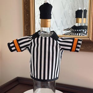 May include: A clear glass wine bottle is dressed in a referee-themed outfit. The bottle wears a black and white striped shirt with orange accents on the sleeves and a black cap. The bottle is displayed on a white surface.