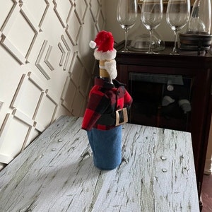 Lumberjack  Santa theme wine bottle cover showing black and red buffalo plaid jacket, blue denim bottoms, red Santa hat,  white beard and thick black belt with gold buckle to complete the look
