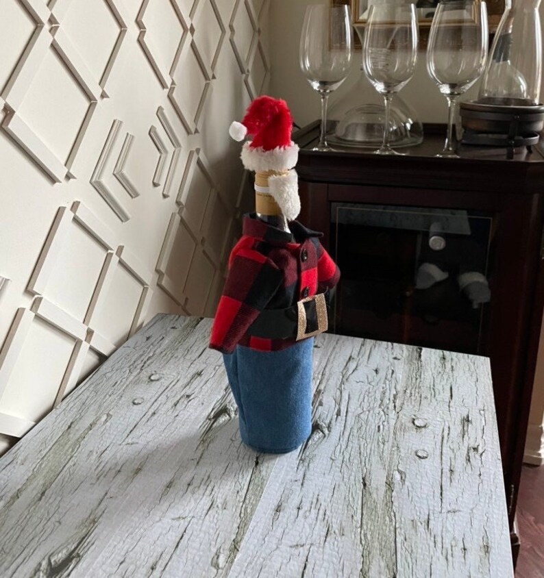 Lumberjack  Santa theme wine bottle cover showing black and red buffalo plaid jacket, blue denim bottoms, red Santa hat,  white beard and thick black belt with gold buckle to complete the look