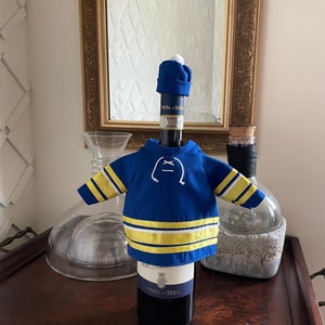 Pode incluir: Uma garrafa de vinho vestida com uma camisa de hóquei azul e amarela com um chapéu branco com pompom. A garrafa é etiquetada com "Tenuta di Sesta" e "DOCG".