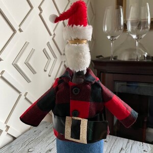 Lumberjack  Santa theme wine bottle cover showing black and red buffalo plaid jacket, blue denim bottoms, red Santa hat,  white beard and thick black belt with gold buckle to complete the look