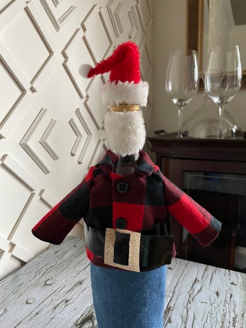 Lumberjack  Santa theme wine bottle cover showing black and red buffalo plaid jacket, blue denim bottoms, red Santa hat,  white beard and thick black belt with gold buckle to complete the look