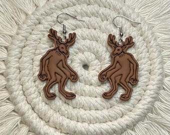 May include: A pair of brown earrings with a stylized humanoid deer design. The earrings have a hook closure and are set against a white, textured background.