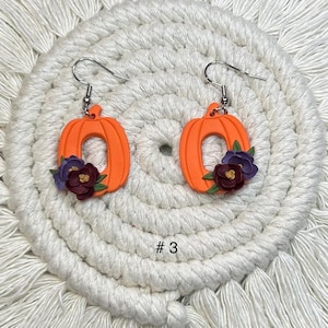 May include: A pair of orange pumpkin earrings with silver hooks. Each earring features a pumpkin shape with a floral arrangement of purple, burgundy, and green accents. The earrings are displayed on a white, textured surface. The number "# 3" is visible.