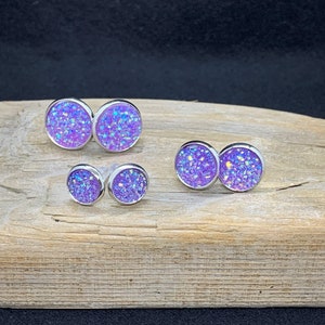 May include: Three pairs of purple glitter stud earrings with silver settings. The earrings are arranged in size order from largest to smallest.