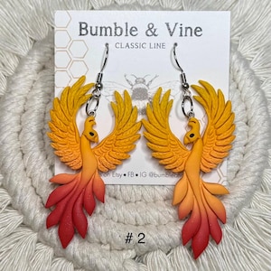 May include: A pair of phoenix earrings in shades of yellow, orange, and red. The earrings are made of a lightweight material and have silver-coloured ear wires. The card behind the earrings has the text "Bumble & Vine CLASSIC LINE" and the number "#2".