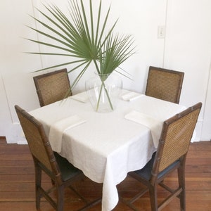 May include: A white tablecloth covers a square wooden table with four chairs. The chairs have woven cane seats and backs. A clear glass vase with a leafy green plant sits in the center of the table.