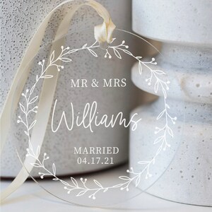 Puede incluir: Un adorno redondo de acr&iacute;lico transparente con un dise&ntilde;o de corona floral blanca. El adorno presenta el texto "MR & MRS Williams MARRIED 04.17.21".